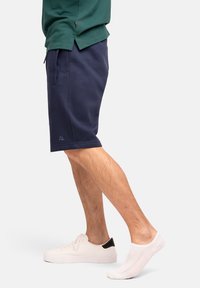Pantalones cortos azul marino de tela suave, con bolsillos laterales y un logotipo, combinados con zapatillas blancas y calcetines blancos cortos.