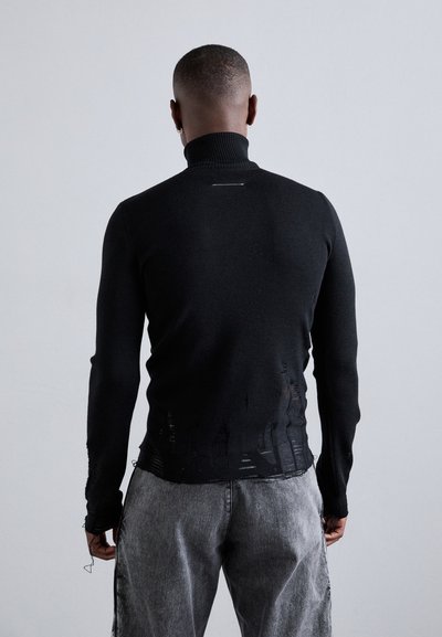 Μαύρο πλεκτό πουλόβερ με ψηλό λαιμό, χαρακτηριστικό άφθαρτο φινίρισμα στο στρίφωμα, μακριά μανίκια και μολυβία σιλουέτα. Φορεμένο με γκρίζα παντελόνια.