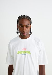 Hvid bomulds t-shirt med et stort grønt grafisk tryk "EXPEDITION" og et lille rødt logo på brystet. Korte ærmer, afslappet pasform.