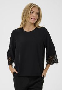 Juoda palaidinė su apvalia iškirpte, ilgomis rankovėmis, kurių gale yra nėrinių detalės, ir laisvo silueto, pagaminta iš minkštos, lengvos medžiagos.