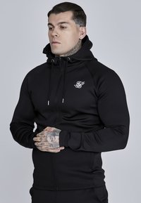 Czarny hoodie zapinany na zamek z troczkami, dopasowany krój, gładka faktura i białe logo na piersi. Model pozuje frontalnie, widoczne tatuaże.
