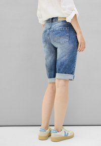 Shorts en denim bleu clair avec un design décoloré, ourlets roulés et poches arrière, associés à des baskets beige avec des accents bleus.