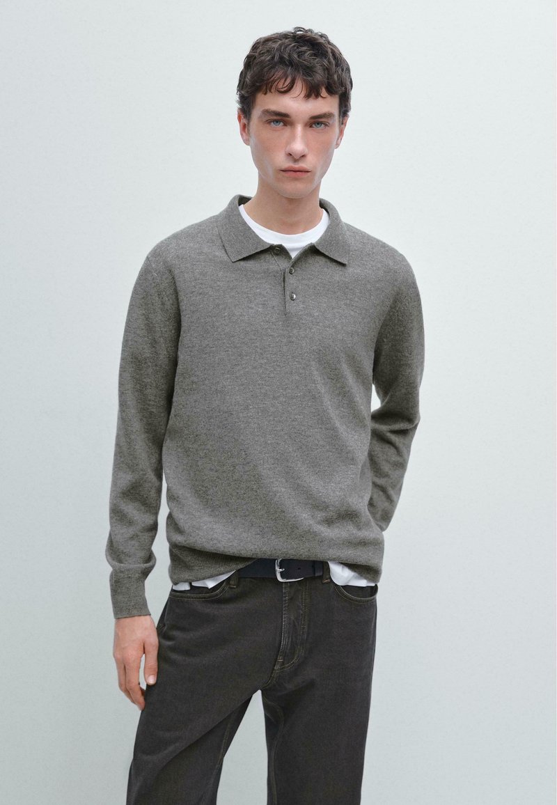 Giovane uomo che indossa un maglione a polo grigio a maniche lunghe sopra una camicia bianca e jeans scuri, in piedi contro uno sfondo chiaro e uniforme.