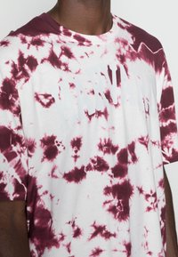Camiseta blanca de manga corta con un patrón de tie-dye en color burdeos, que presenta un audaz texto en blanco "HARVARD" en el pecho.