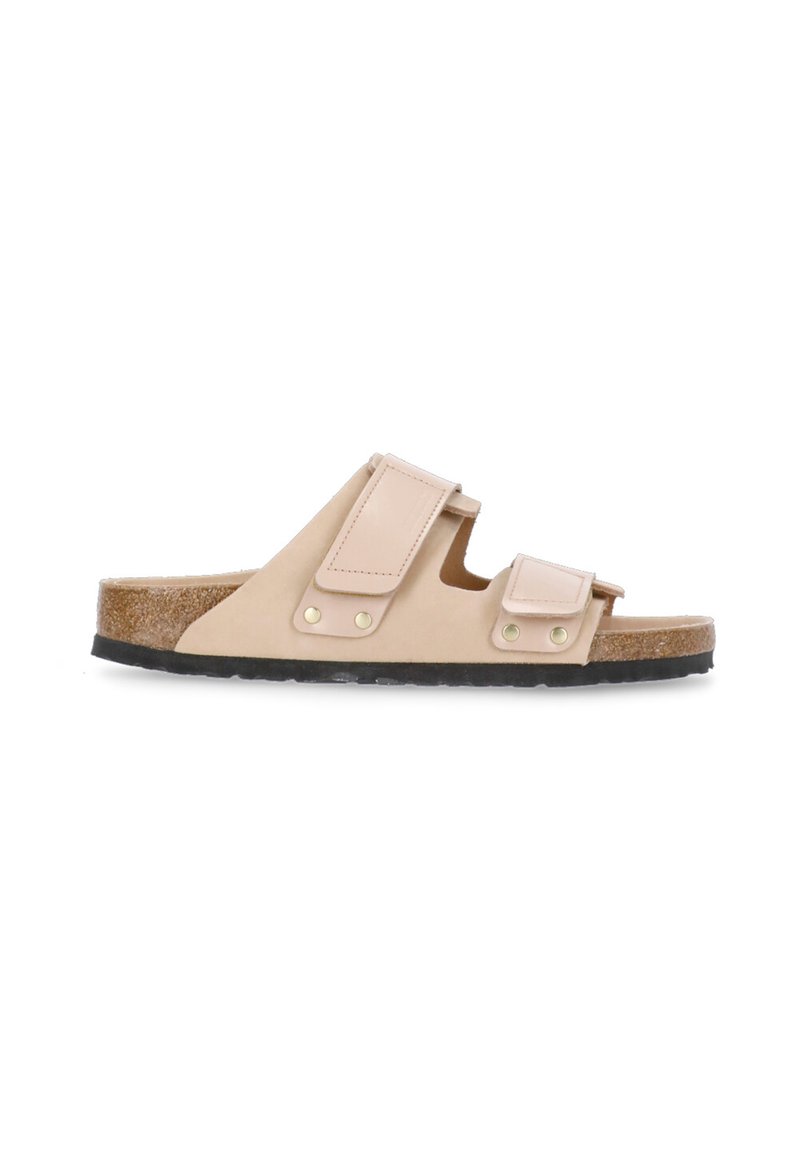Sandalia deslizante beige con dos correas ajustables, acentos dorados, plantilla de corcho y suela de goma negra, con una textura suave.