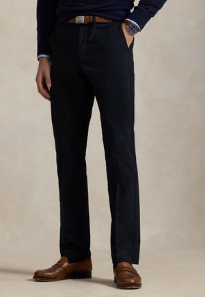 Homme portant un pantalon noir, une ceinture en cuir marron, des mocassins marron, un pull bleu marine et une montre-bracelet sur un fond neutre.
