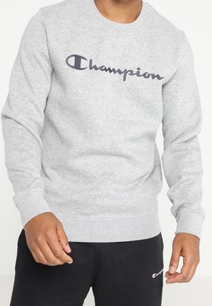 Man draagt een lichtgrijze Champion trui en zwarte Champion joggingbroek, staat met ontspannen handen langs zijn zij tegen een effen achtergrond.