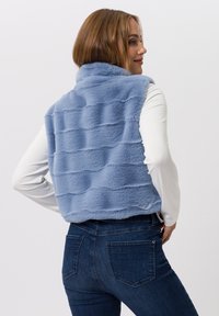 Hellblauer, plüschiger Kunstpelz-Weste mit einer gerippten Textur und hohem Kragen, kombiniert mit einem taillierten weißen Langarmshirt und dunklen Jeans aus Denim.