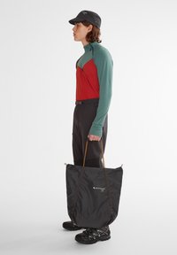 Schwarze Tasche mit Kordelzug, auf der ein Logo zu sehen ist. Modell trägt ein rot-grünes Langarmhemd und schwarze Hose und hält die Tasche.
