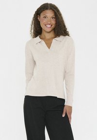 Light beige long-sleeve top with a collar, featuring a relaxed fit and soft texture. Worn with dark pants, set against a plain background.