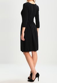 Donna che indossa un abito nero lungo fino al ginocchio con maniche a tre quarti e scarpe nere con tacchi alti, in piedi di lato su uno sfondo bianco.