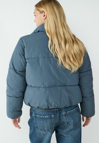 Kurtka puchowa w odcieniu szarym, z wysokim kołnierzem, przeszytymi panelami i krojem krótkim. W zestawieniu z niebieskimi dżinsami.