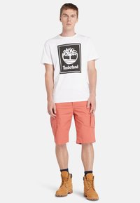 Balta trumpa rankovė T-marškiniai su juodu Timberland logotipu, derinami su oranžiniais krovinių šortais ir šviesiai rudos spalvos lacai batais.