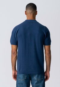 Mørkeblå kortærmet polo-skjorte lavet af tekstureret stof, med en flad krave og en lige kant, parret med lyseblå denim jeans.