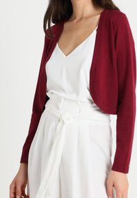 Burgundy cropped cardigan made of soft fabric, featuring a round neckline and open front, layered over a white top and high-waisted trousers.