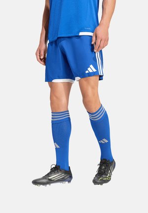 Mężczyzna sportowiec ubrany w niebieskie spodenki piłkarskie, dopasowane skarpetki z białymi paskami oraz czarne korki, stojący na tle jednolitego tła.