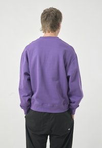 Purple sweatshirt with a crew neck, long sleeves, and a relaxed fit. Made of soft fabric, featuring a ribbed hem and cuffs. Black pants visible.