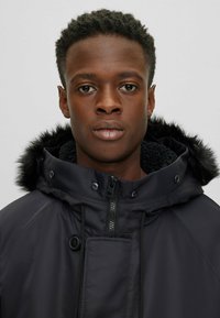 Parka negra con un alto cuello, capucha con pelo y cierre frontal de cremallera. Presenta un forro interior texturizado y cuerdas de ajuste.