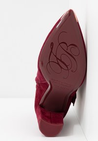 Scarpa con tacco alto in suede bordeaux con punta affusolata e un fiocco decorativo sul davanti. Suola liscia e lucida con dettagli del marchio impresso.