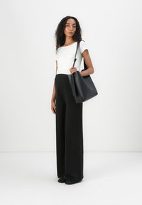 Svarte vide bukser kombinert med en hvit tettsittende topp. Modellen holder en stor svart skinn totebag. Bakgrunnen er helt hvit.