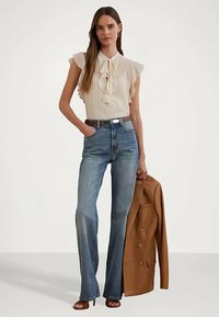 Frau in einer cremefarbenen Rüschbluse, blauen High-Waist-Jeans, braunem Gürtel, die ein braunes Blazerjacke hält und braune Absatzsandalen trägt.