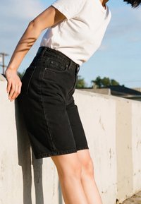 Personne portant un short noir jusqu'aux genoux et un t-shirt blanc, adossée à un mur en béton blanc en extérieur.