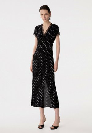 Donna che indossa un abito midi nero con pois bianchi, rifinitura in pizzo sullo scollo e sulle maniche e spacco frontale, abbinato a tacchi neri.