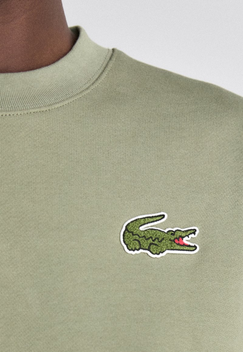 Felpa verde chiaro con collo a costine, con un logo di coccodrillo verde ricamato sul lato sinistro del petto con dettagli rossi e neri.