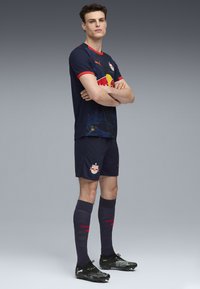 Equipación de fútbol azul oscura con un acento rojo en el cuello, que presenta un logo. Botas negras y calcetas azules oscuras con detalles en texto rojo.