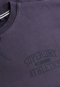 Fioletowa bluza z prążkowanym kołnierzem i mankietami, ozdobiona ciemnym nadrukiem logo "Superdry Athletics" na przodzie.