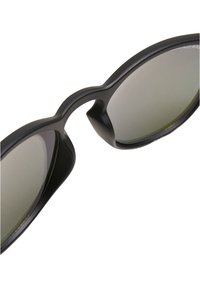 Lunettes de soleil noires avec une finition lisse et mate. Monture incurvée et verres teintés de vert. Détail de pic notable entre les verres.