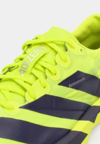 Αθλητικό παπούτσι νέον κίτρινο με μαύρο ριγέ σχέδιο, υφή στην επάνω πλευρά και κορδόνια. Διαθέτει λογότυπο στη γλώσσα και αεριζόμενο υλικό.