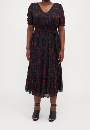 Vrouw die een zwarte midi-jurk draagt met rood bloemenprint, V-hals, korte pofmouwen, gelaagde rok, tailleband en zwarte open tenen enkellaarzen met bandje.