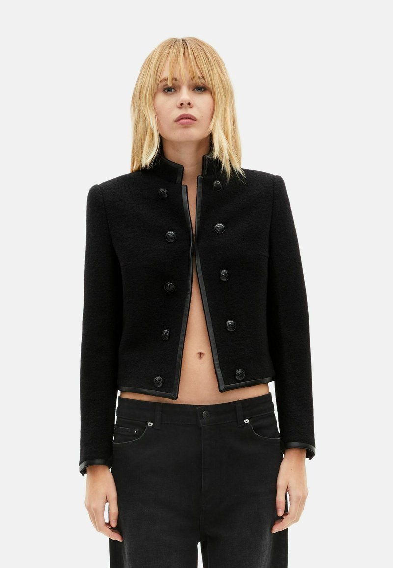 Giacca corta nera con colletto alto, tessuto texturizzato, chiusura frontale con bottoni e rifiniture in pelle nera. Abbinata a pantaloni in denim nero.