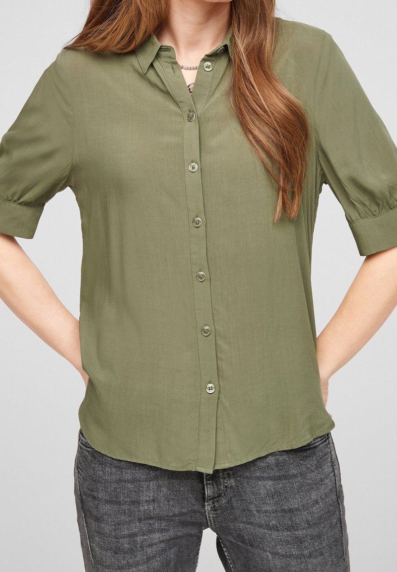 Femme portant une chemise kaki à manches courtes avec col et boutons, associée à un jean gris foncé, debout les mains sur les hanches devant un fond gris.
