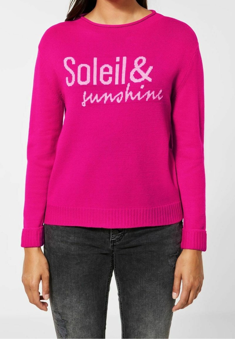Pull rose vif à manches longues avec le texte "Soleil & sunshine" en rose clair, porté avec un jean gris foncé sur une personne montrée du cou aux hanches.