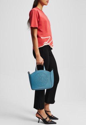 Light blue leather handbag with perforated round design, carried by a person wearing a coral shirt and black trousers with a split hem.