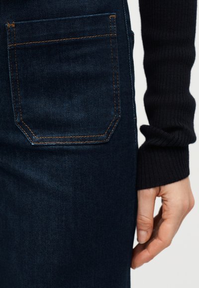 Κοντινή λήψη πίσω τσέπης σκούρου μπλε τζιν με πορτοκαλί ραφές, δίπλα σε ένα χέρι που ακουμπά κοντά στο ισχίο φορώντας μαύρο πλεκτό μανίκι.