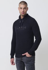 Marineblaues Strickhoodie mit strukturiertem Muster, das einen hohen Kragen und Kordelzüge aufweist. Das Design beinhaltet ein dezentes Logo über der Brust.