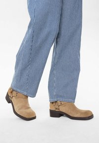 Bottines en suède beige à bout arrondi, petit talon carré et sangle décorative avec des clous métalliques ; portées avec un pantalon rayé bleu et blanc.