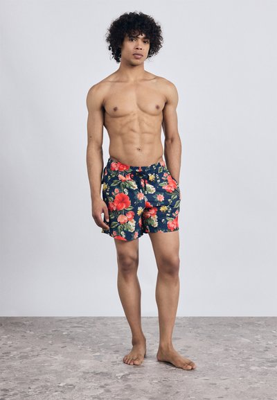 Jeune homme debout pieds nus sur un sol en pierre, portant un short de bain à motifs floraux avec les mains dans les poches, contre un fond gris uni.