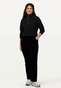 Zwarte ritstrui met ribbelstructuur, gecombineerd met zwarte corduroy broek en witte sneakers, met een relaxte pasvorm en zakken.