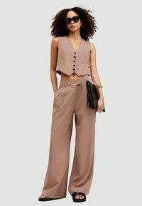 Woman wearing beige sleeveless buttoned vest and wide-legged trousers, holding black clutch, with curly hair and black sunglasses.