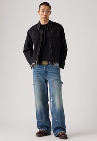 Chaqueta bomber negra con tachuelas metálicas, camiseta negra, jeans de pierna ancha de color azul claro con bolsillos, zapatos de cuero marrón, hebilla de cinturón plateada.