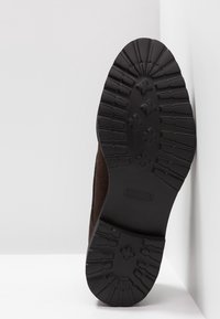 Bruine suède schoen met een gestructureerde zwarte rubberen zool, voorzien van een geprofileerde loop voor grip en een gebogen ontwerp. Zool rechtop weergegeven.