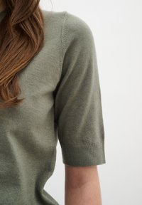Maglione corto a maniche corte di un verde oliva chiaro con una texture morbida, scollatura rotonda e polsini a coste. Cuciture decorative sulle spalle.