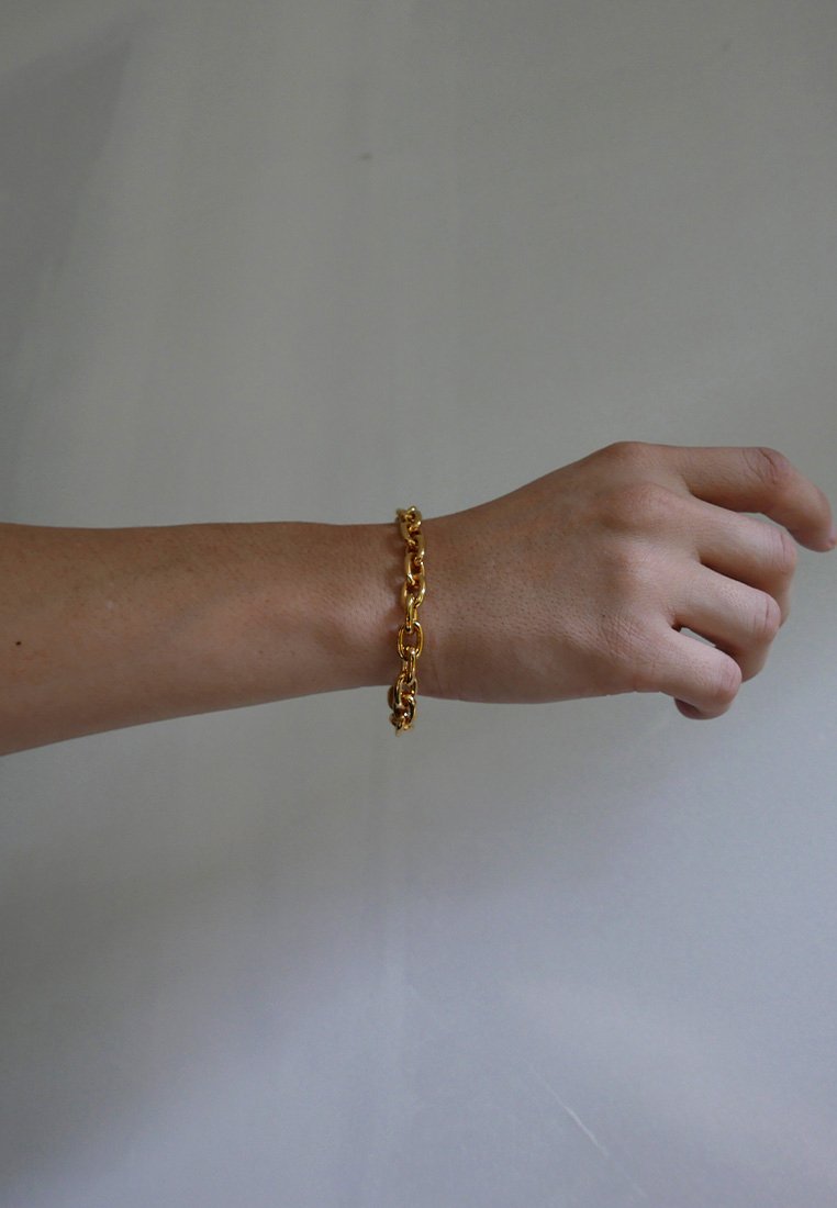 Pulsera de cadena en tono dorado con eslabones entrelazados, superficie suave, acabado pulido y un cierre seguro, llevada en una muñeca sobre un fondo neutro.