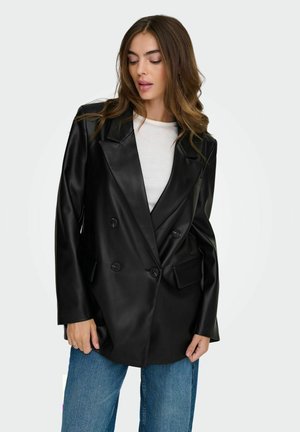 Femme portant un blazer noir à double boutonnage en faux cuir sur une chemise blanche et un jean bleu, regardant vers le bas avec les cheveux détachés.