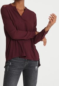 Blusa bordeaux realizzata in tessuto leggero, con scollatura a V, maniche lunghe e dettagli in pizzo sulle spalle. Abbinata a jeans neri strappati.
