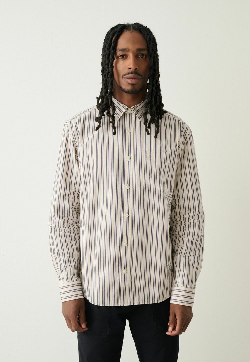 Košile na knoflíky s vertikálními pruhy v krémové, námořnické a tmavě hnědé barvě. Má límec s špičkami a jedno náprsní kapsu. Bavlněná látka.